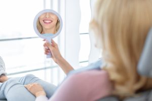 Woman admiring her bright smile in a handheld mirror in a dental office.