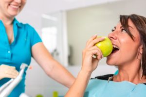 Patient biting a green apple while smiling with dentist in blue scrubs observing.